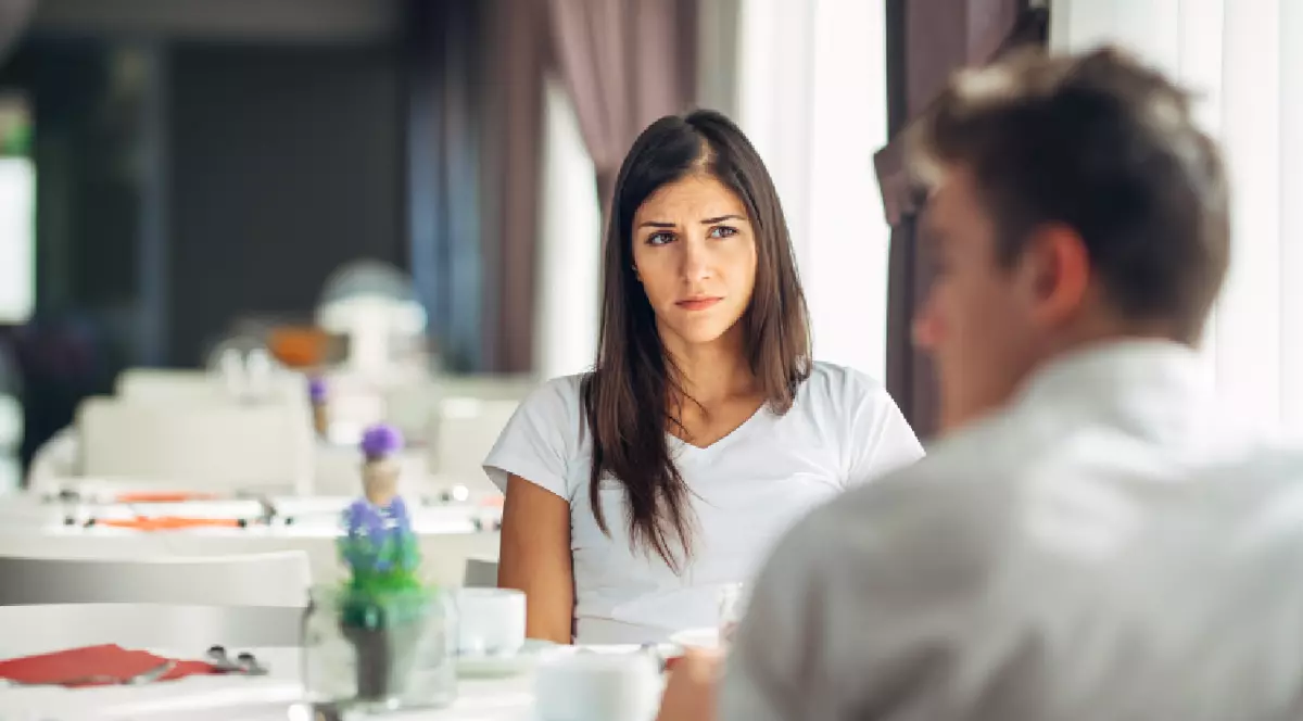 O femeie și un bărbat, îmbrăcați în alb, stau la o masă, într-un restaurant gol