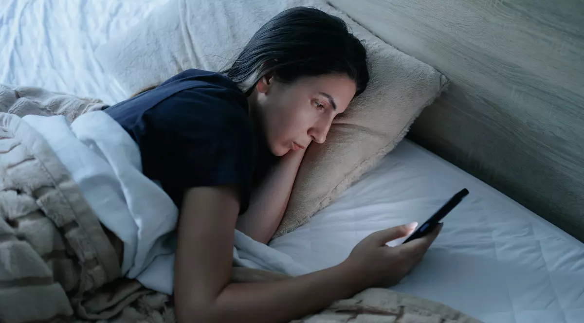 O femeie care stă în pat cu capul pe perna și cu telefonul în mână, în timp ce privește în ecran