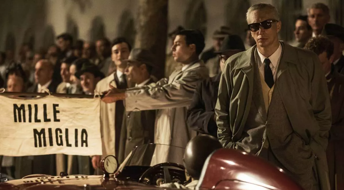 Scena din filmul FERRARI, 2023, se vede Adam Driver in prim plan, în rolul Enzo Ferrari, in spatele unei masini Ferrari rosie, in spate o multime de oameni, figuranti