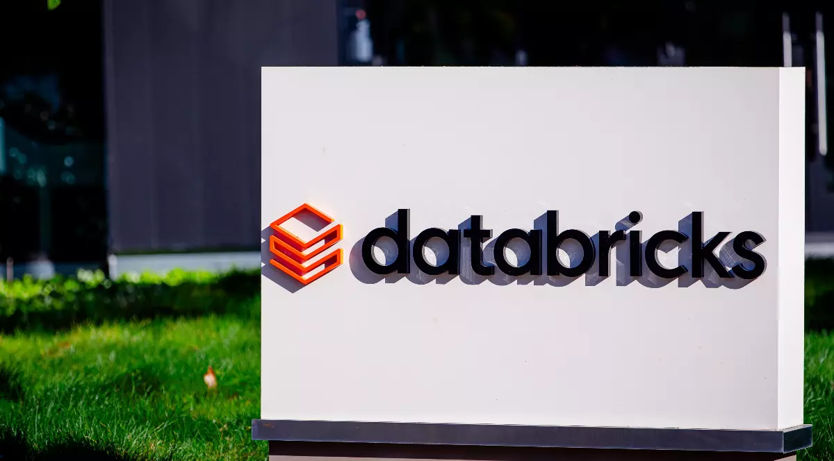 Imaginea prezintă un panou alb amplasat în fața unei clădiri moderne, pe care este afișat logo-ul companiei Databricks, alături de simbolul său portocaliu stilizat. Panoul este amplasat pe o peluză verde, sugerând intrarea într-un campus sau sediu corporativ al companiei.