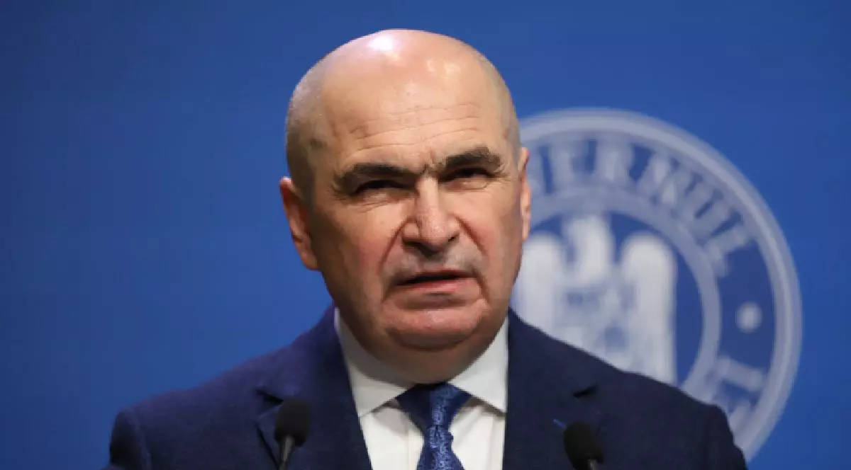 Imaginea îl prezintă pe Ilie Bolojan, îmbrăcat într-un costum bleumarin și cămașă albă, vorbind în fața unor microfoane pe fundalul unui panou oficial albastru ce conține o stemă.