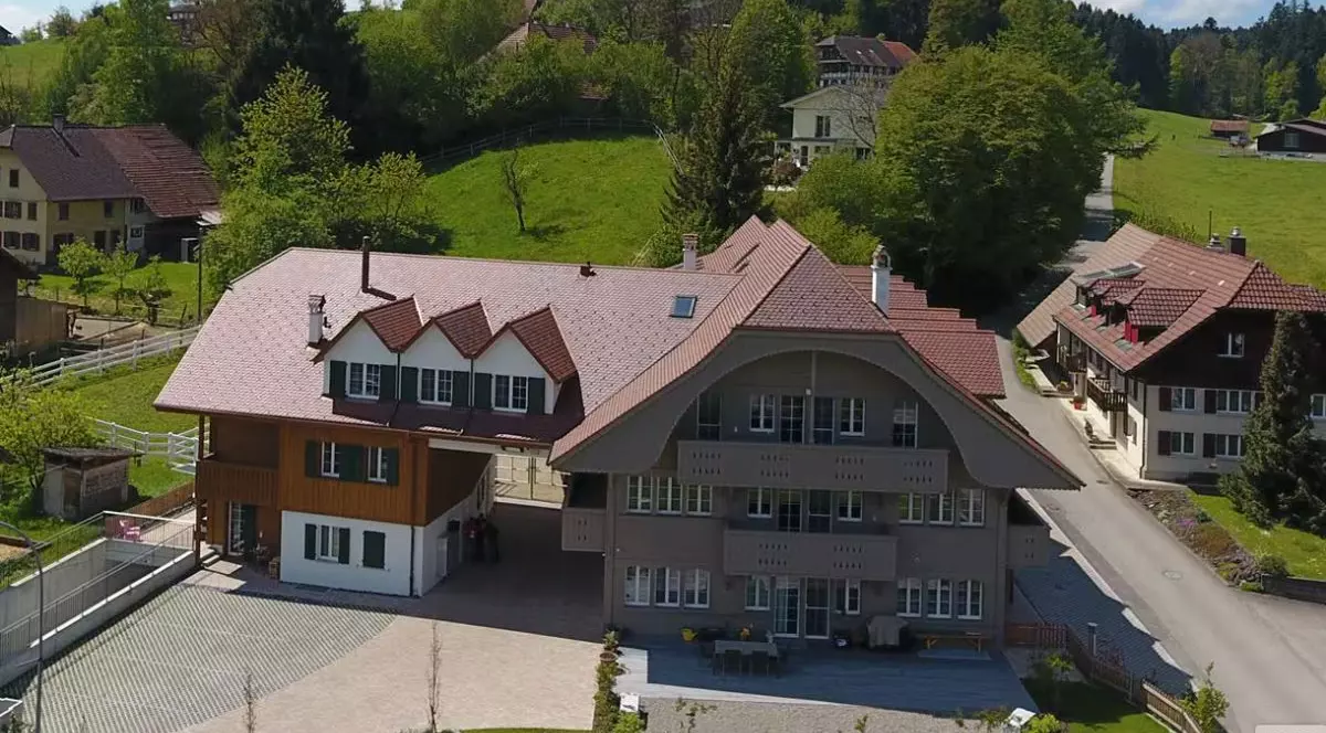 Clădirea fermei, cu acoperiș de culoare maro închis, înconjurată de pădure și pășune