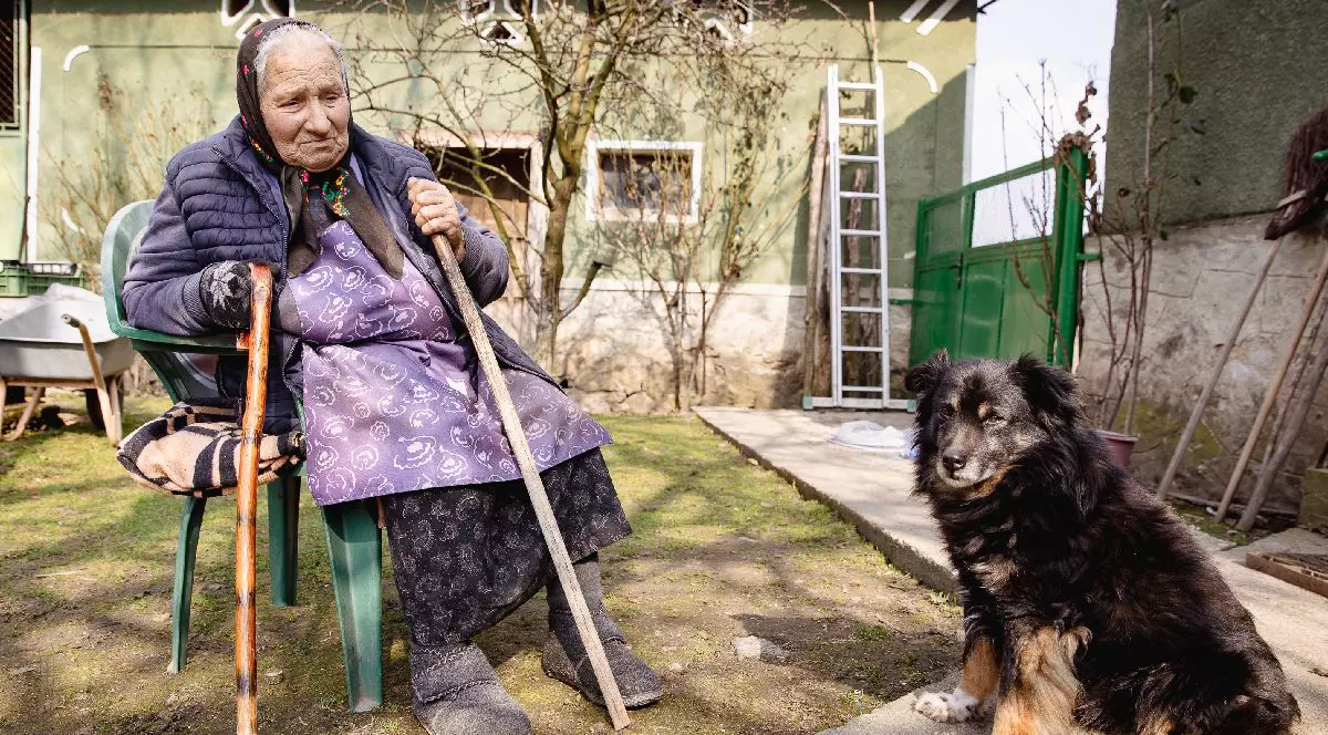 Floarea Bolojan, o femeie în vârstă, mama premierului Ilie Bolojan, stând pe un scaun în curtea casei ei din Birtin, iar alături un câine negru