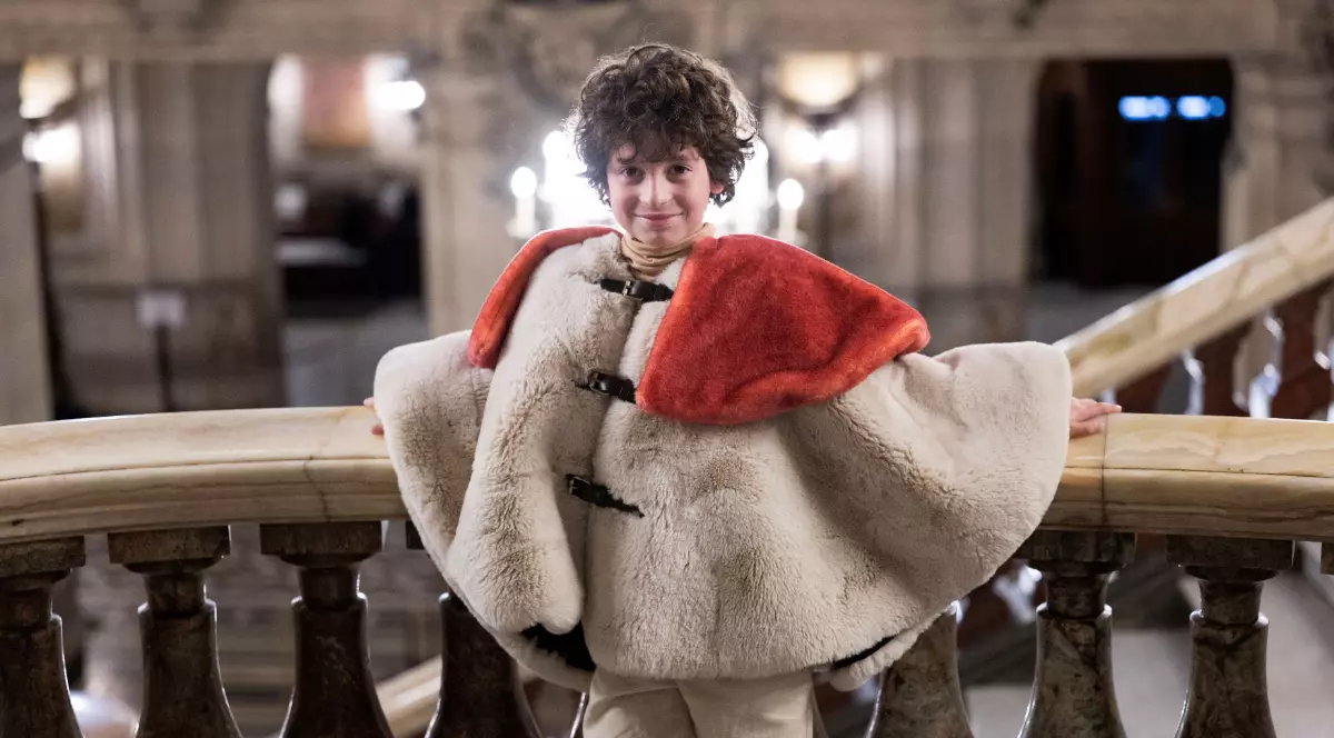 Un copil stă sprijinit de balustrada unei scări monumentale într-o clădire elegantă, purtând o piesă vestimentară voluminoasă din blană deschisă la culoare, cu un guler roșu. Ținuta pare parte dintr-o colecție de modă, iar decorul somptuos sugerează atmosfera unui eveniment sau a unei prezentări într-un loc prestigios.