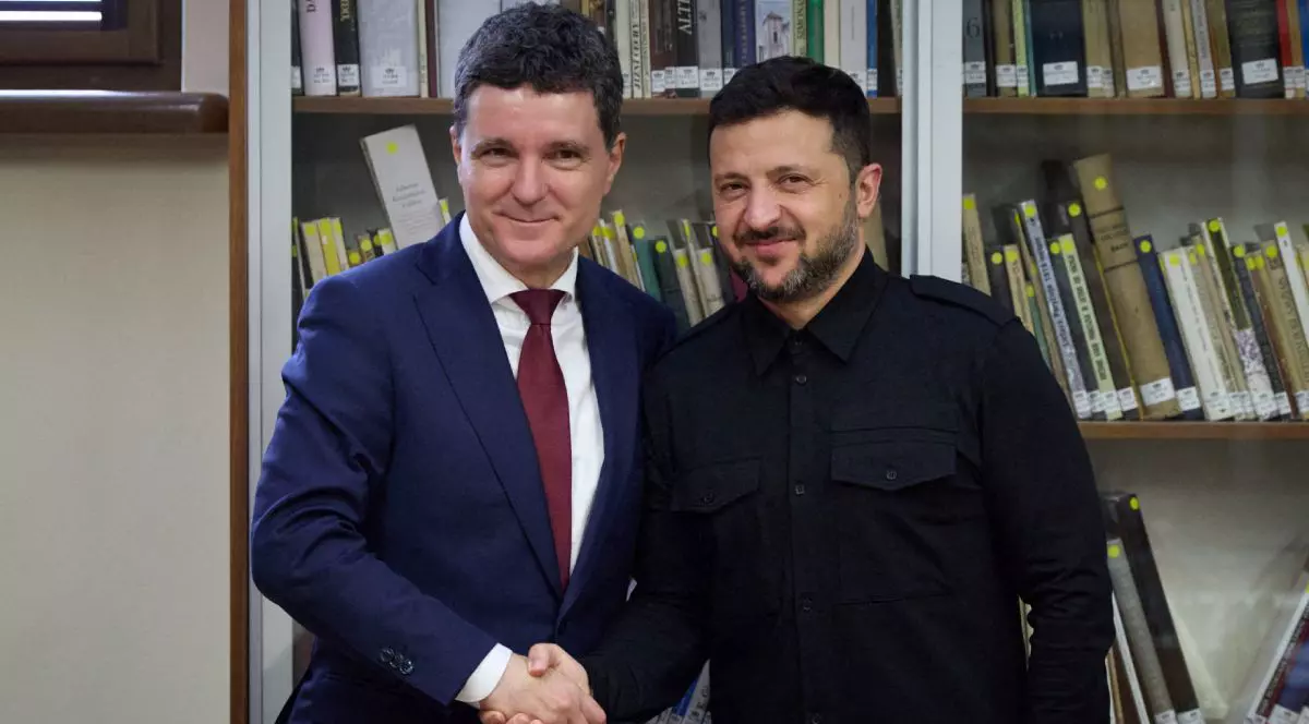 Nicușor Dan, îmbrăcat în costum albastru, cămașă albă și cravată roșie și Volodimir Zelenski, când dau noroc, îmbrăcat cu o bluză neagră, iar pe fundal o bibliotecă plină cu cărți