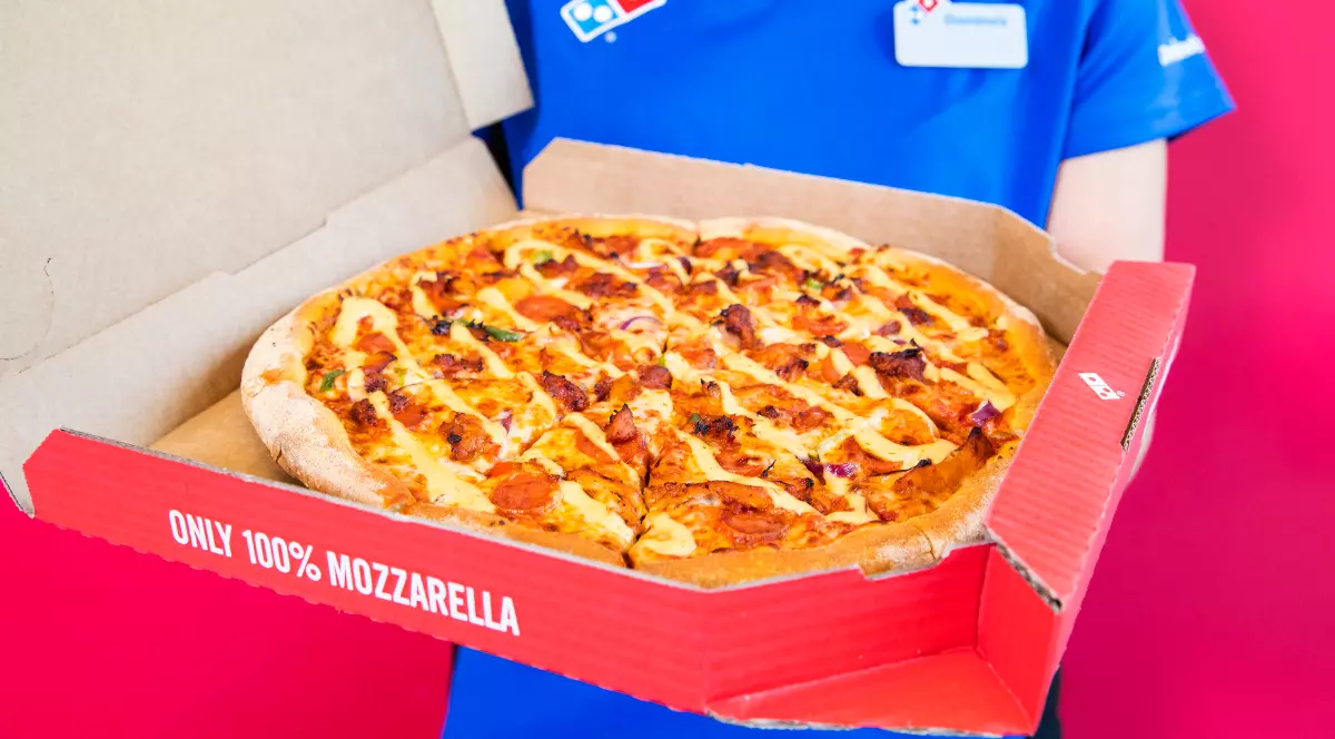 O persoană îmbrăcată în uniforma albastră Domino’s ține o cutie deschisă cu o pizza proaspăt preparată, cu brânză topită și diverse toppinguri. Fundalul roșu intens și logo-ul companiei evidențiază imaginea specifică unei livrări de fast-food.