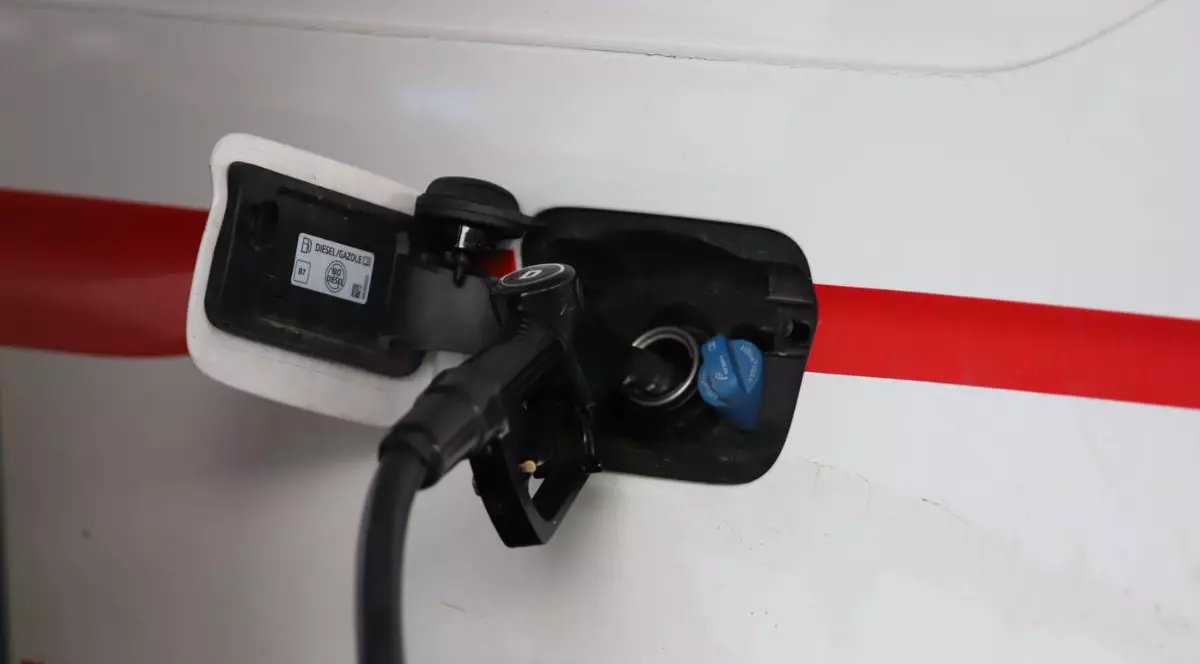 prim-plan al zonei de alimentare a unui vehicul de culoare albă, prevăzut cu o bandă orizontală roșie pe caroserie. Capacul rezervorului este deschis, lăsând la vedere interiorul unde un pistol de alimentare negru este introdus în orificiul principal. Pe partea interioară a capacului alb se află un autocolant cu specificații tehnice, unde sunt vizibile textele „DIESEL/GAZOLE” și simbolul „B7”.