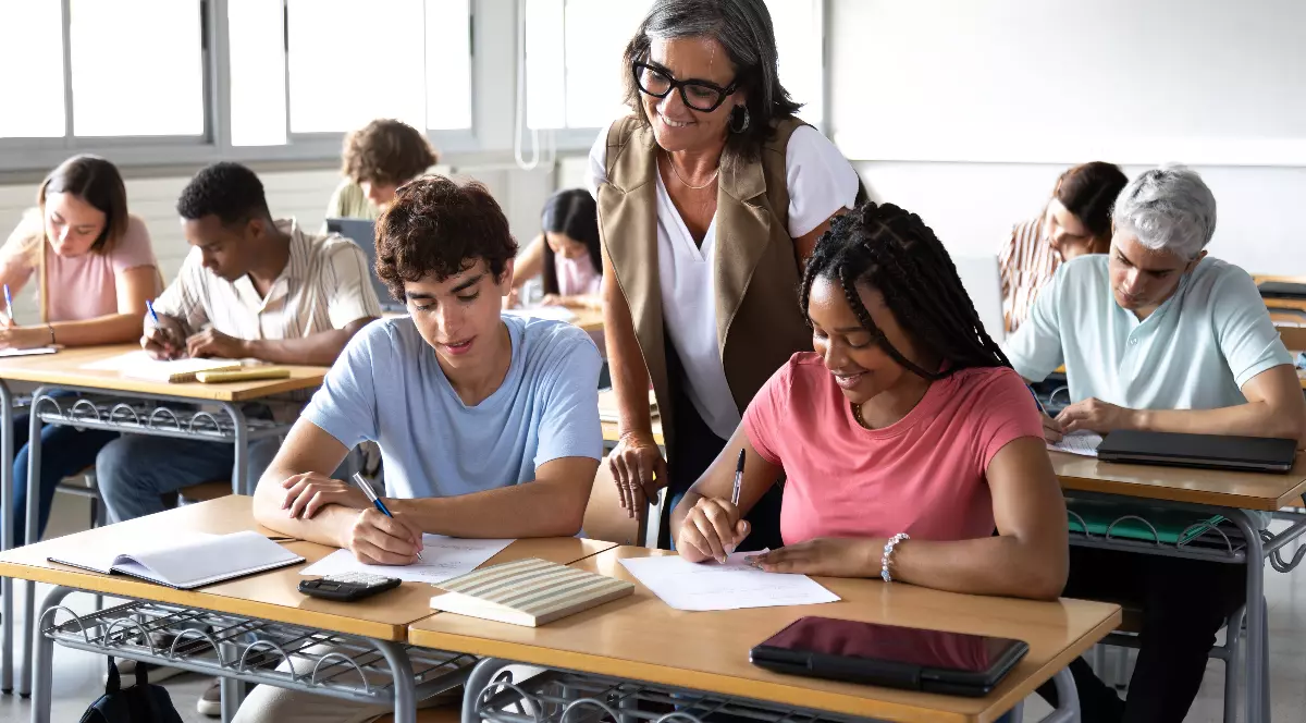 O profesoară îi asistă pe elevi într-o sală de clasă luminoasă, în timp ce aceștia scriu la teste sau exerciții la bănci. Atmosfera este una concentrată și calmă, sugerând un moment obișnuit de activitate școlară.