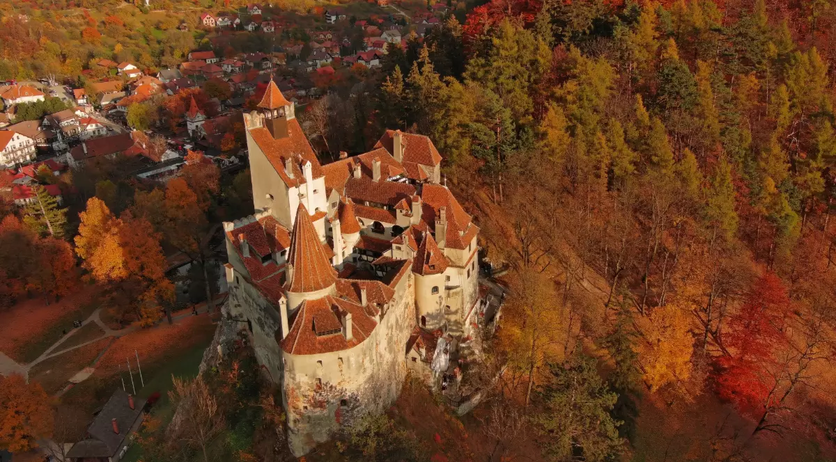 Imaginea surprinde Castelul Bran, așezat pe o stâncă și înconjurat de o pădure colorată în nuanțele toamnei. În fundal se vede localitatea din apropiere, cu case și străzi care completează peisajul montan spectaculos.
