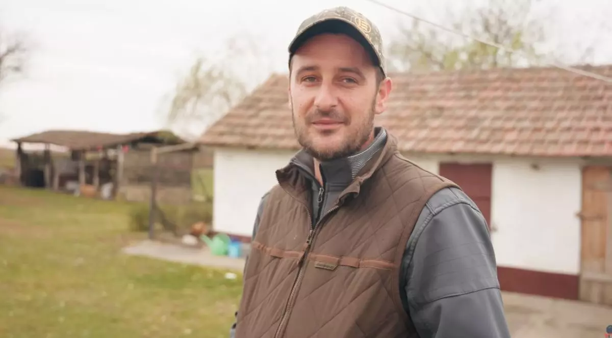 Imaginea surprinde un tânăr care privește direct spre cameră și zâmbește ușor. Poartă o șapcă de camuflaj, o vestă maro peste o jachetă gri și se află într-o zonă rurală. În fundal se vede o casă tradițională cu pereți albi și acoperiș din țiglă.