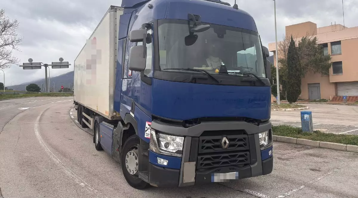 Fotografie cu un camion albastru mare cu remorcă albă, parcat pe o stradă, într-o zi înnorată, cu clădiri și indicatoare rutiere în fundal.