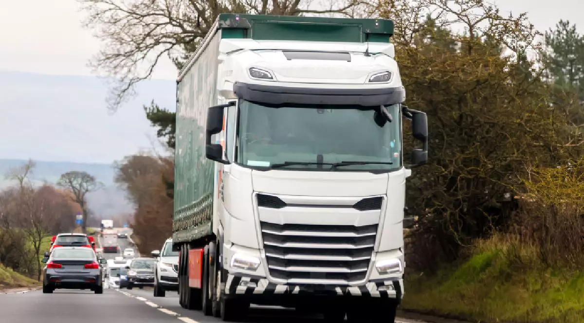 Fotografie cu un camion alb cu remorcă verde care circulă pe un drum cu două benzi, înconjurat de copaci, cu mai multe mașini în fundal.