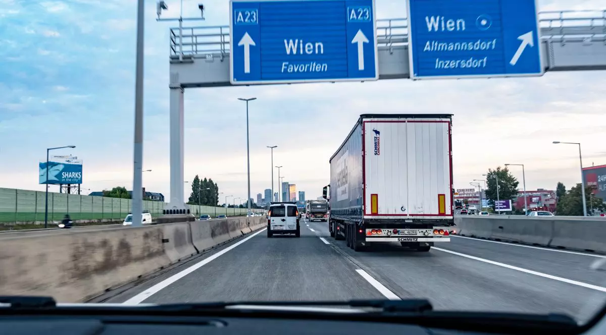 Fotografie cu o autostradă aglomerată, cu un camion alb și o dubă albă în prim-plan, semne rutiere albastre deasupra și un cer senin în fundal.