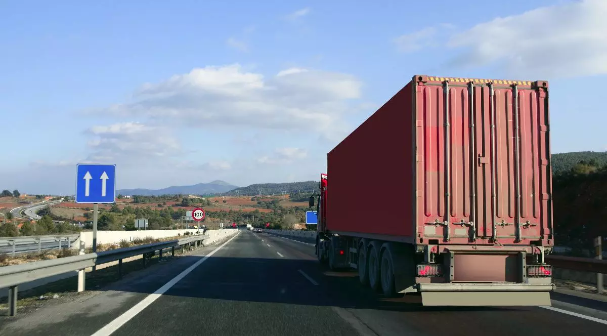 Un TIR cu remorcă roșie circulă pe un drum cu mai multe benzi, sub un cer senin, traversând un peisaj deluros.
