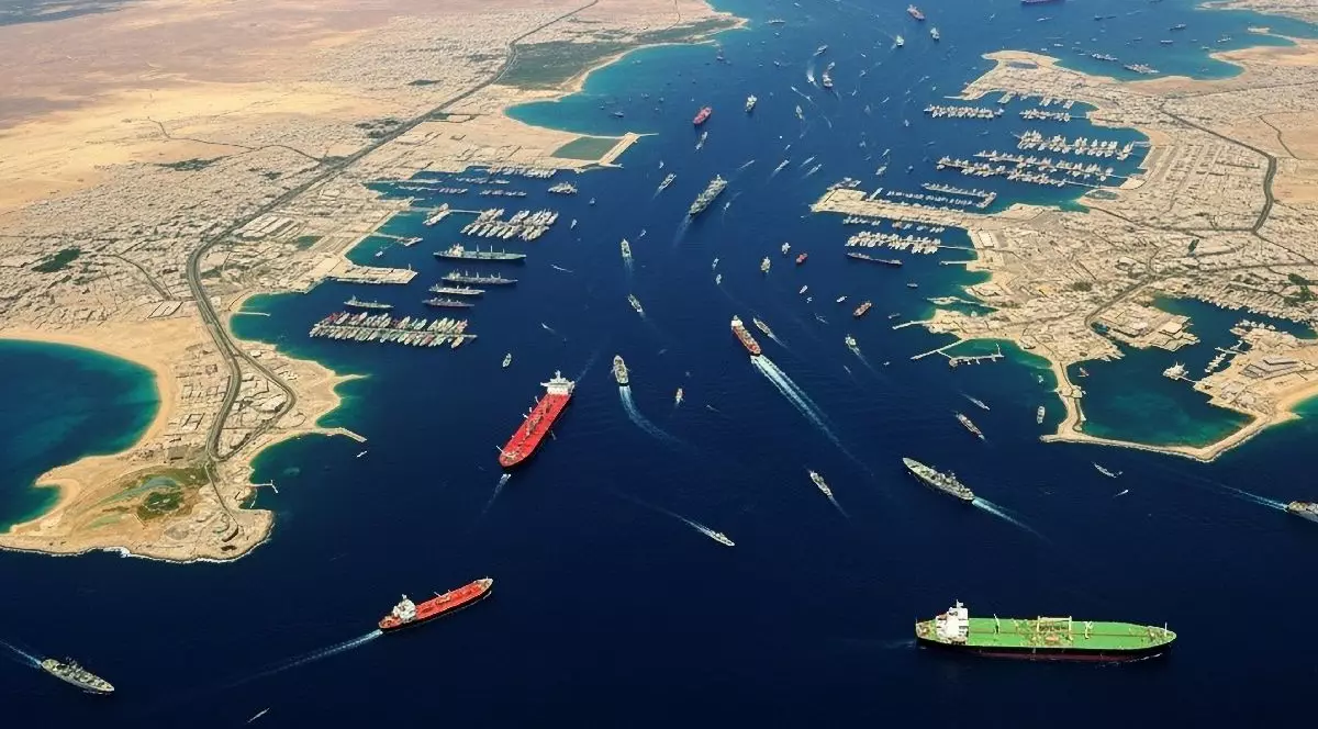 Imaginea prezintă o vedere aeriană a unei căi navigabile. Numeroase nave de dimensiuni mari, inclusiv cargouri, tancuri petroliere și nave militare, tranzitează canalul.