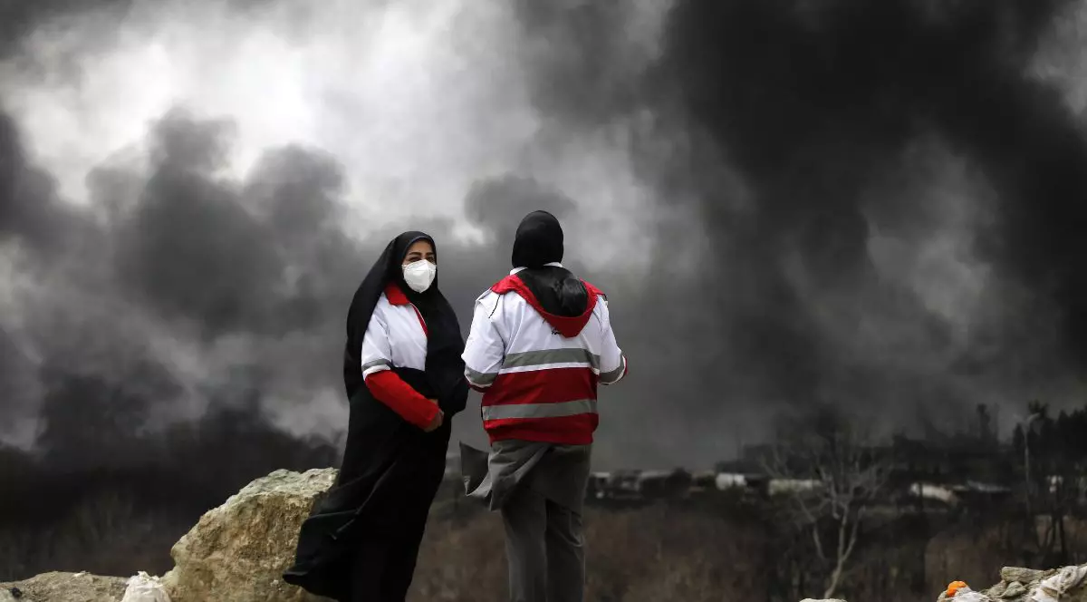 Această fotografie prezintă două persoane care stau pe un teren accidentat, acoperit cu roci și dărâmături, pe un fundal dominat de nori groși și denși de fum gri închis și negru care acoperă aproape întregul cer. Persoana din stânga, orientată parțial spre dreapta, poartă un acoperământ negru pe cap, o mască de protecție albă și o jachetă albă cu umeri și mâneci roșii peste haine negre. Persoana din dreapta este surprinsă din spate, purtând o jachetă similară, albă cu roșu și cu glugă, și un acoperământ negru pe cap. În plan secund, sub stratul masiv de fum, se disting contururile neclare ale unor structuri sau vehicule îndepărtate și vegetație rară, lipsită de frunze, Teheran, 8 martie 2026