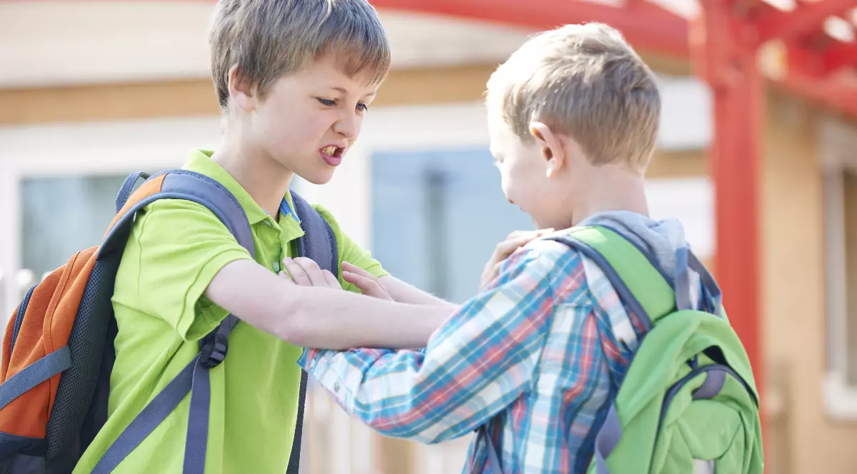 Doi băieți, cu ghiozdane în spate, par să se certe sau să se împingă în apropierea unei școli. Unul dintre ei este vizibil nervos, sugerând un conflict între copii.
