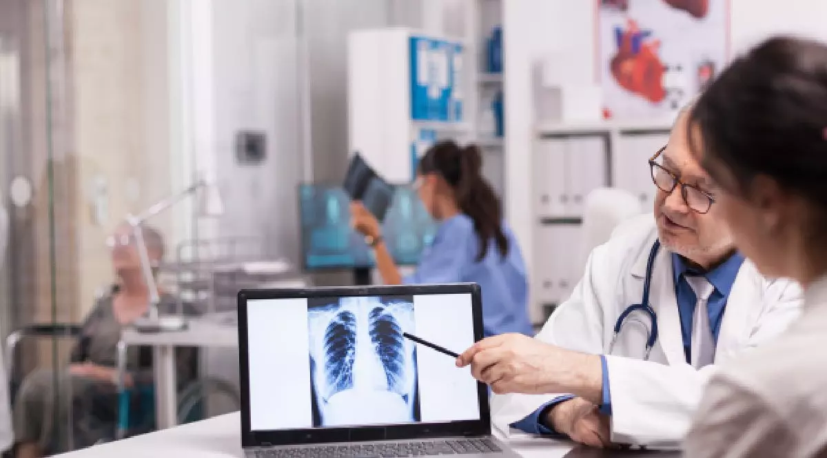 Intr-un cabinet medical, o femeie in halat albastru tine o radiografie in mana, un alt cadru medical in halat alb si cu stestoscop la gat arata cu batul spre o radiografie pulmonara. Tuberculoza in Romania