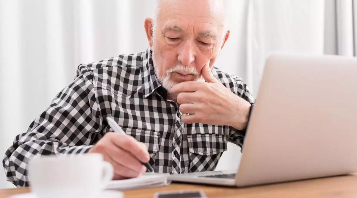 Un bărbat cu părul alb este la birou în fața unui laptop folosind și scrie într-un carnețel. Pe masă mai sunt cafeaua și un telefon mobil