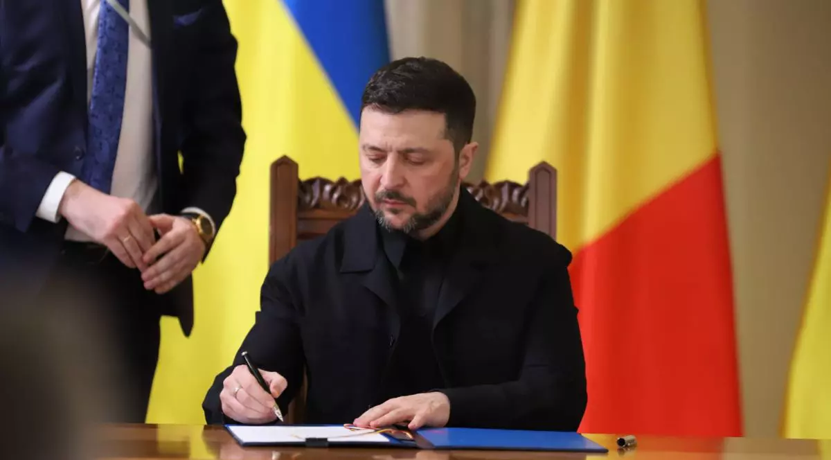 Volodimir Zelenski semnând un act, îmbrăcat în negru, cu tricolorul pe fundal