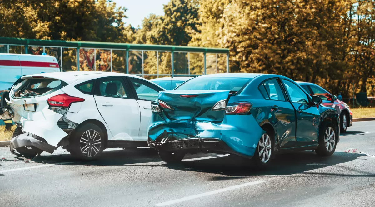 Un accident între două mașini, cea din stânga albă, iar cea din dreapta, albastră. Autoturismele s-au cionit lateral, pe o șosea. În jurul lor, copaci, vara.