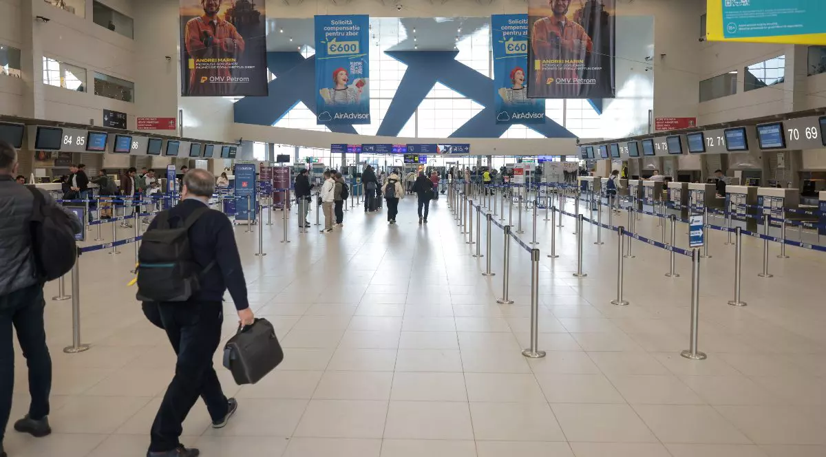 Imaginea prezintă interiorul unui aeroport, cu mai mulți pasageri care se deplasează spre ghișeele de check-in. Spațiul este modern, bine organizat, cu zone delimitate pentru fluxul de călători și panouri informative suspendate.