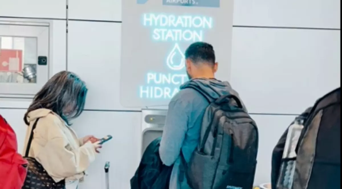 Imaginea surprinde un punct de hidratare instalat în interiorul unui aeroport, unde pasagerii își pot umple sticlele cu apă. În fața stației, mai multe persoane așteaptă sau își verifică telefoanele, în timp ce folosesc această facilitate.