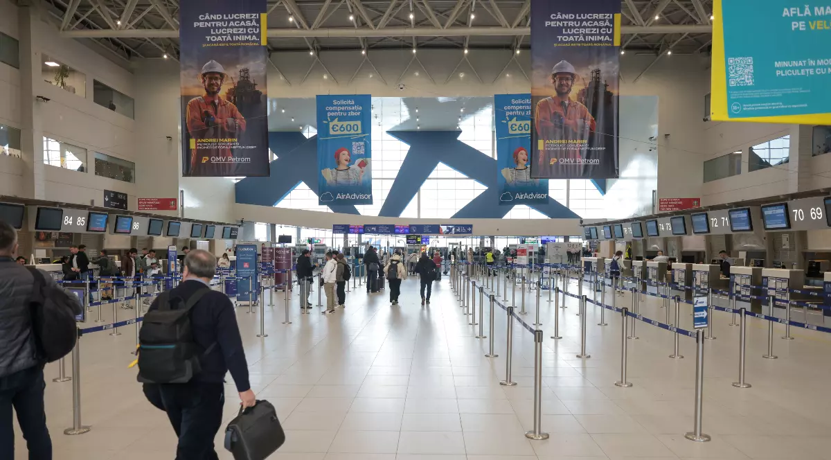 Imaginea prezintă interiorul unui aeroport, cu mai mulți pasageri care se deplasează spre ghișeele de check-in. Spațiul este modern, bine organizat, cu zone delimitate pentru fluxul de călători și panouri informative suspendate.