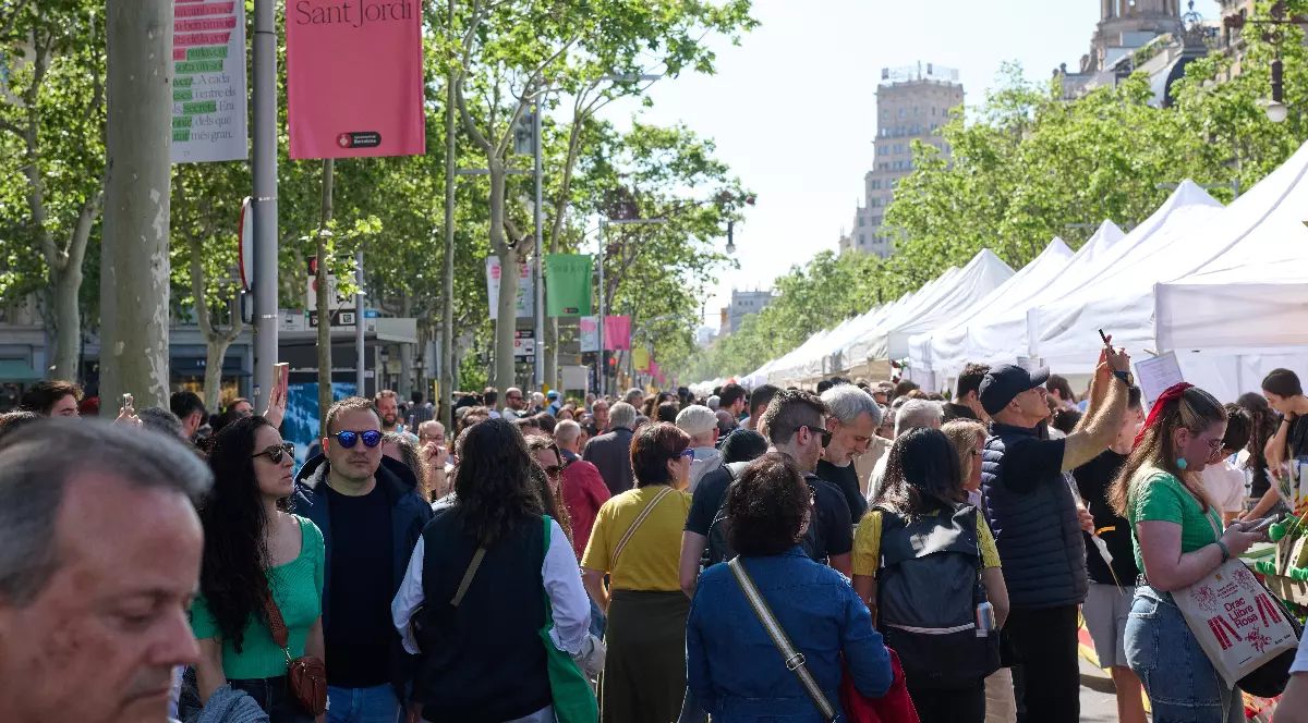 Imaginea surprinde o mulțime numeroasă de oameni adunați pe o stradă largă, unde are loc un eveniment sau târg în aer liber. De-o parte și de alta se văd standuri acoperite și bannere colorate, iar atmosfera este animată și aglomerată.