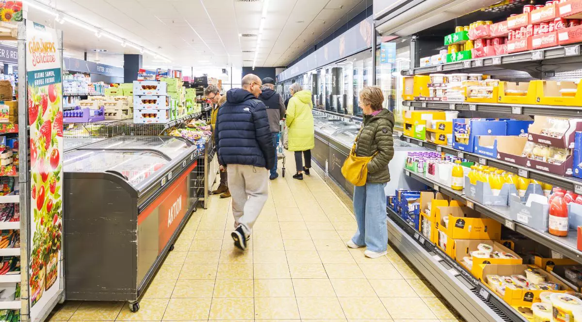 Imaginea prezintă mai mulți clienți care fac cumpărături pe culoarul unui supermarket, examinând produsele din vitrinele frigorifice și de pe rafturile pline cu alimente și băuturi.