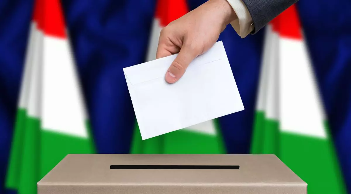 mana unui bărbat în sacou gri, sub care e vizibilă manșeta unei cămăși albe, introducând un vot într-o urnă, cu steagul ungariei pe fundal - alegeri parlamentare ungaria 12 aprilie 2026