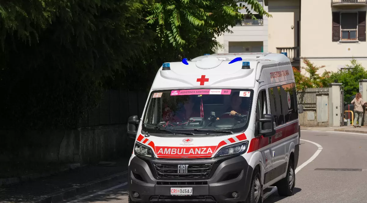 Imaginea prezintă o ambulanță văzută printr-un efect de suprapunere sau reflexie, cu luminile albastre aprinse, sugerând o intervenție de urgență. În fundal se observă siluete de persoane și elemente distorsionate, ceea ce creează o atmosferă tensionată și dramatică.