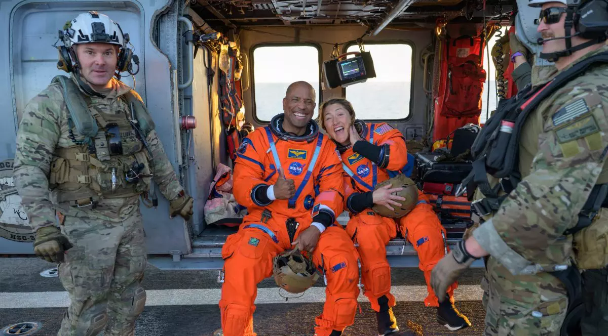 Jumătate din echipajul misiunii Artemis II stă pe marginea unui elicopter, cu soldați alături.