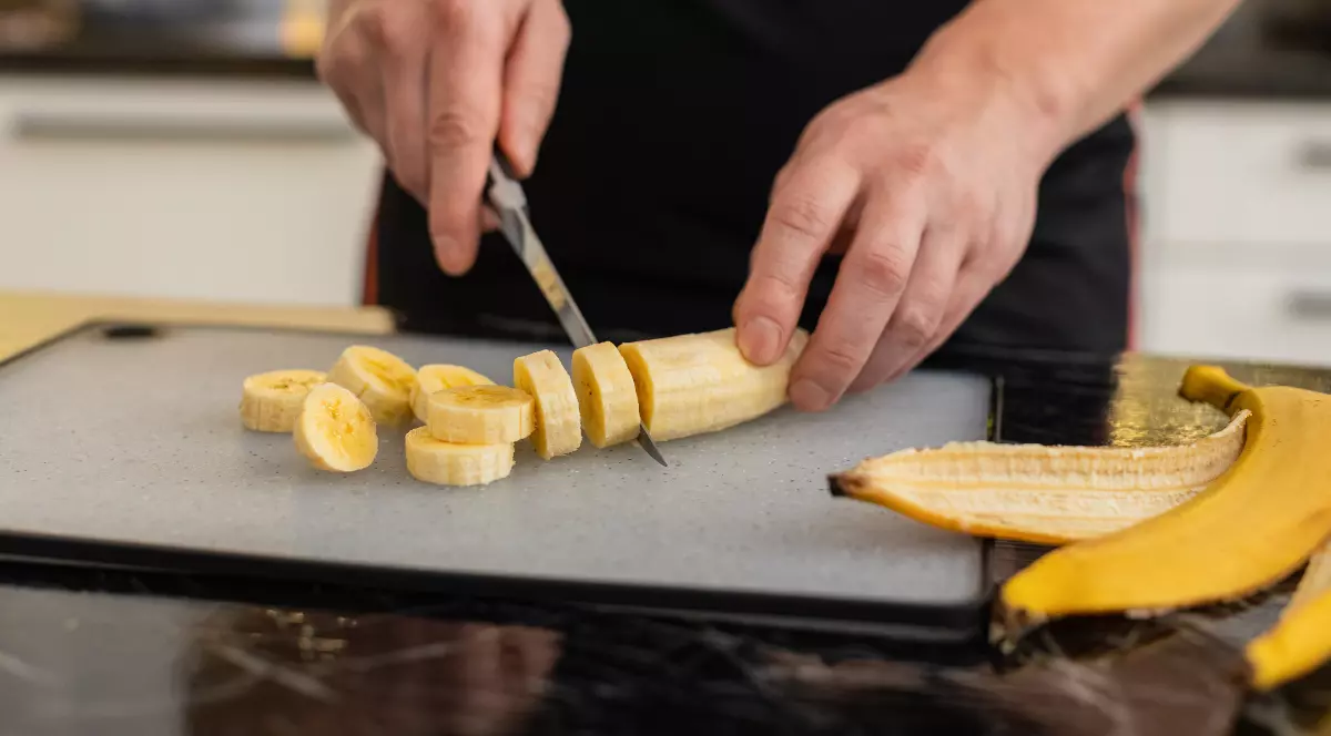 Imaginea arată o banană tăiată felii pe un tocător, lângă ambalaje de produse alimentare. În prim-plan se vede un cuțit care taie banana, sugerând pregătirea unui aliment sau a unei gustări.