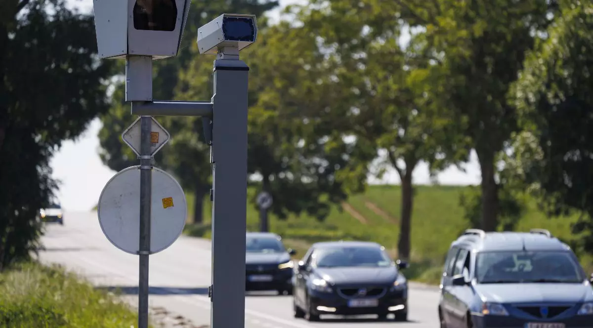 Imaginea surprinde două camere de supraveghere a traficului montate pe un stâlp metalic pe marginea unui drum, monitorizând autoturismele care circulă printr-o zonă rurală cu vegetație bogată.