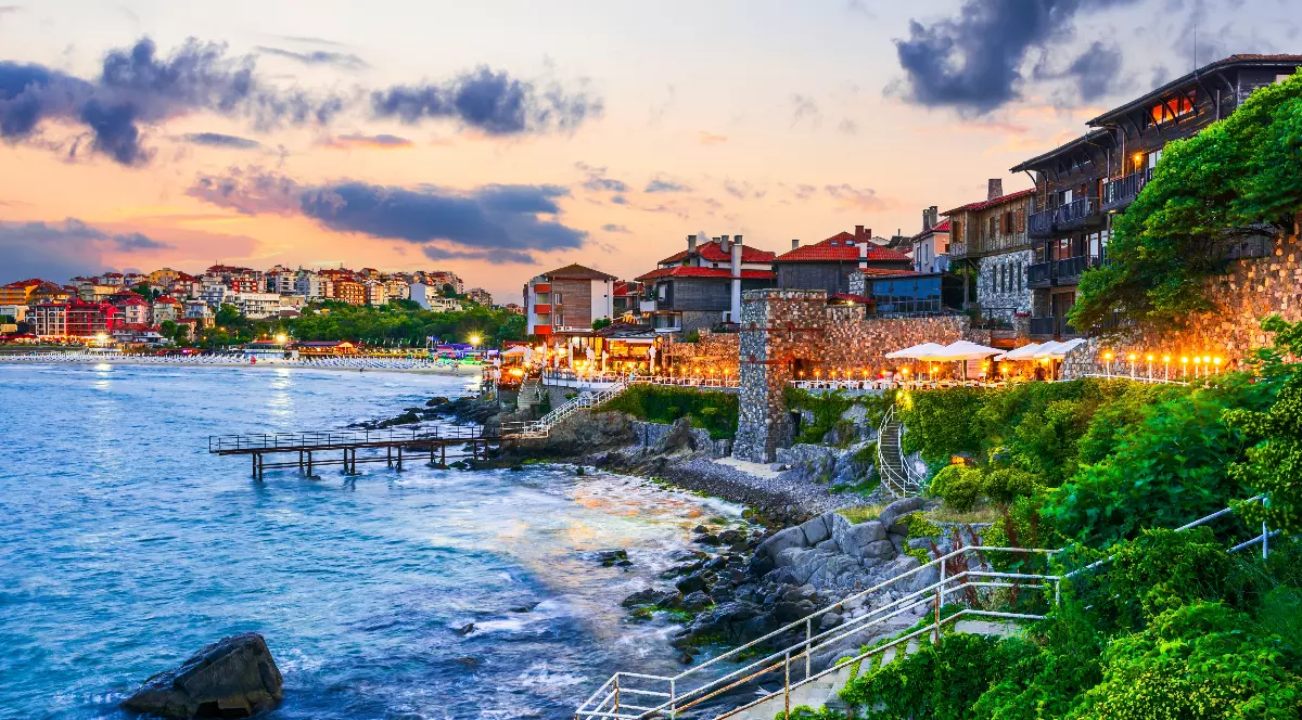 Imaginea surprinde o stațiune de pe litoralul bulgăresc, cu case și restaurante amplasate pe malul mării, luminate cald la apus. Valurile se sparg de stânci, iar atmosfera este relaxantă și pitorească, sugerând o destinație de vacanță atractivă.