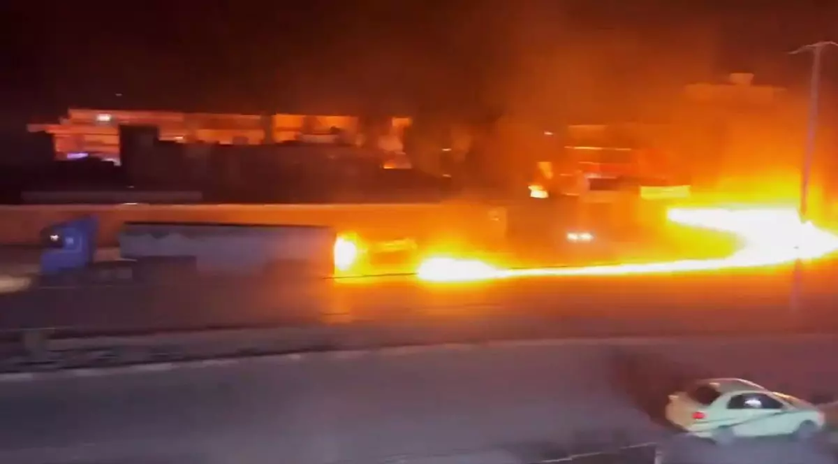 Imaginea surprinde o scenă nocturnă dramatică în care un incendiu violent se propagă sub formă de râu de foc de-a lungul unei șosele, în apropierea unui camion.