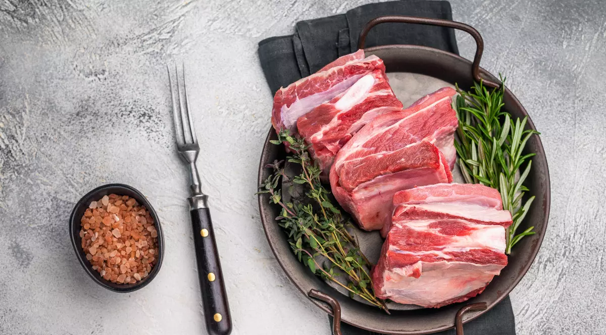Mai multe bucăți de carne de miel crudă sunt așezate într-o tavă, alături de ierburi aromatice precum rozmarinul și cimbrul. În apropiere se află sare grunjoasă și o furculiță, sugerând pregătirea pentru gătit.