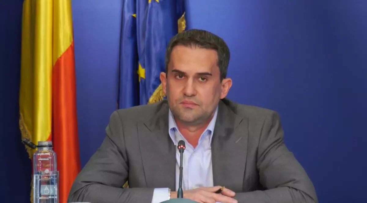 Un bărbat îmbrăcat la costum stă la o masă în timpul unei conferințe de presă, având în față un microfon. În spatele său se află steagul României și cel al Uniunii Europene, sugerând un context oficial.