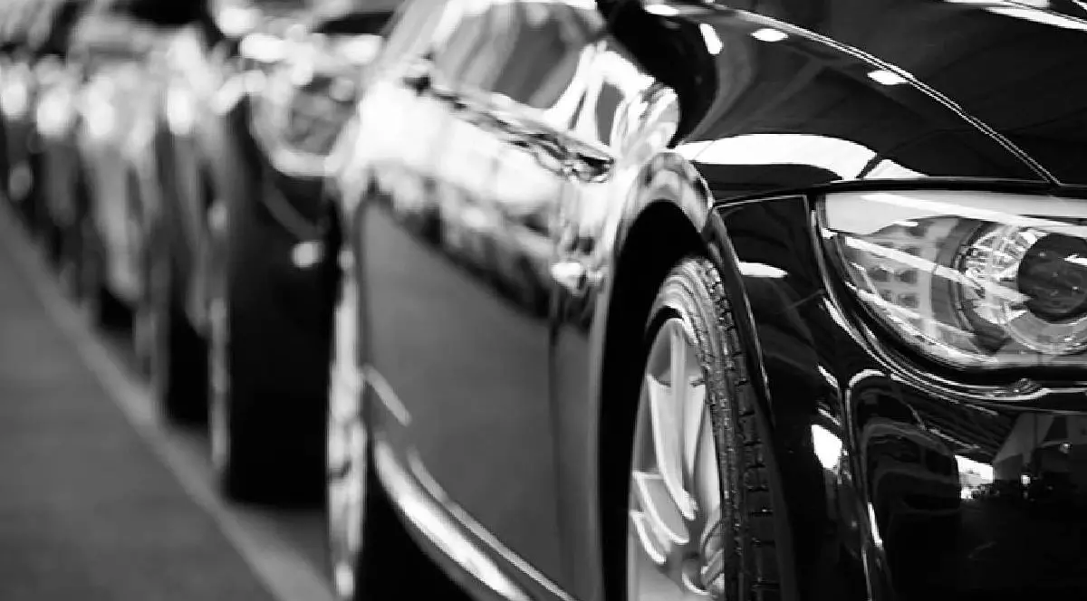 Imagine alb-negru cu mai multe mașini parcate în linie, surprinse din lateral. În prim-plan se vede clar roata și farul unei mașini, în timp ce restul vehiculelor sunt ușor neclare în fundal, sugerând un șir de automobile aliniate.