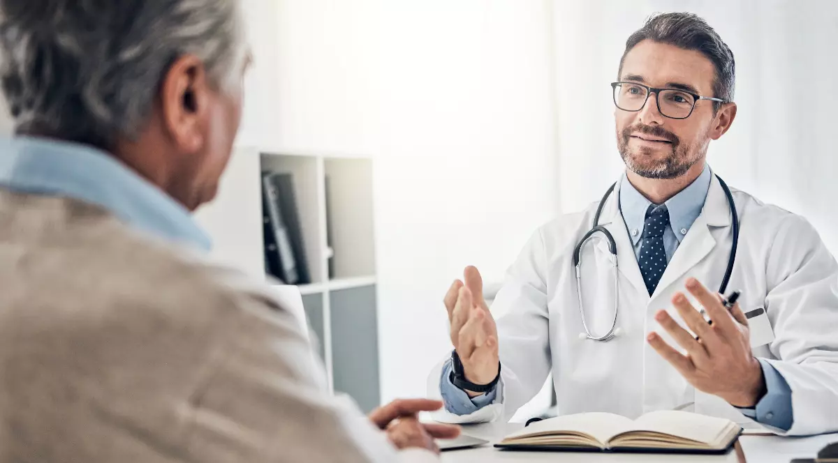 Imaginea surprinde un medic în timpul unei consultații, discutând cu un pacient aflat în fața sa. Doctorul pare atent și explică detalii, într-un cadru medical modern și luminos.