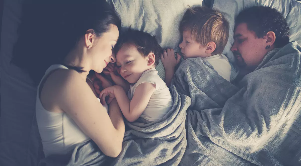 O familie doarme împreună în același pat, într-o atmosferă liniștită și caldă, sugerând apropiere și siguranță. Cei doi copii sunt între părinți, acoperiți cu o pătură, iar toți par relaxați și odihniți.