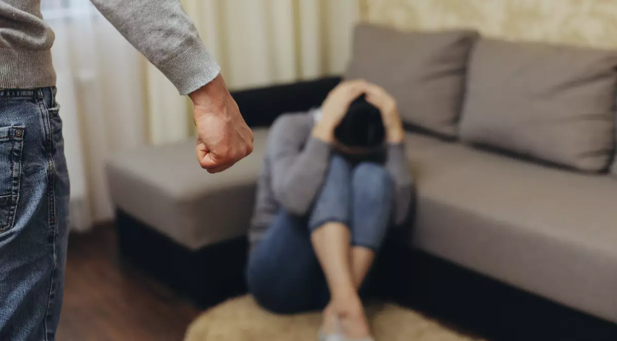 O femeie care stă jos, cu mâinile pe cap. În partea stângă este un bărbat care ține pumnul strâns.