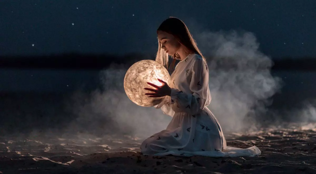 Imagine creativă pe timp de noapte, cu o femeie îmbrăcată în rochie lungă, care stă în genunchi și ține în mână un glob luminat din interior