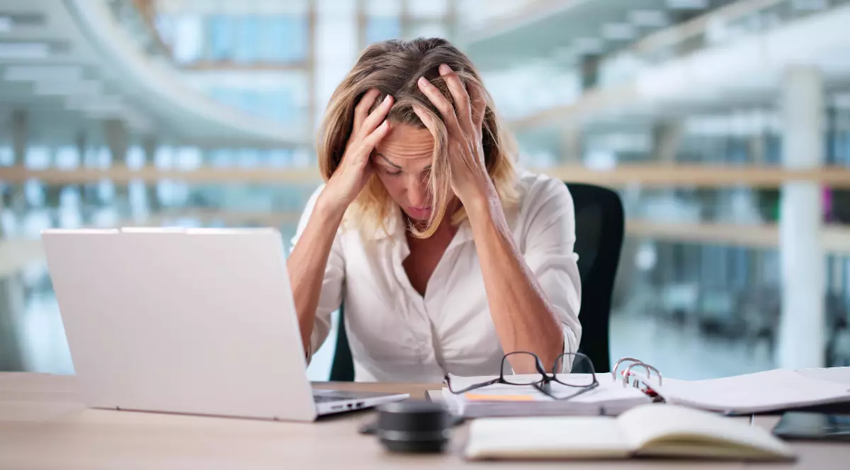 O femeie blondă de peste 50 de ani care stă la birou cu mâinile în cap