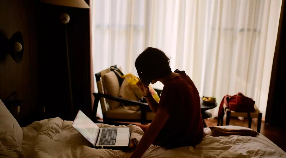 Persoană așezată pe un pat folosind un laptop într-o cameră slab luminată, cu perdele transparente.