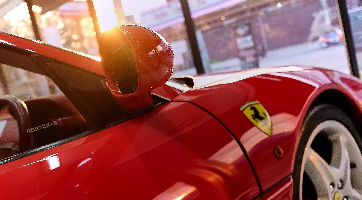 Imaginea prezintă un detaliu de prim-plan al unui autoturism Ferrari roșu strălucitor, evidențiind emblema celebră cu căluțul cabrat și oglinda laterală, totul fiind scăldat în lumina apusului care se reflectă pe caroserie.