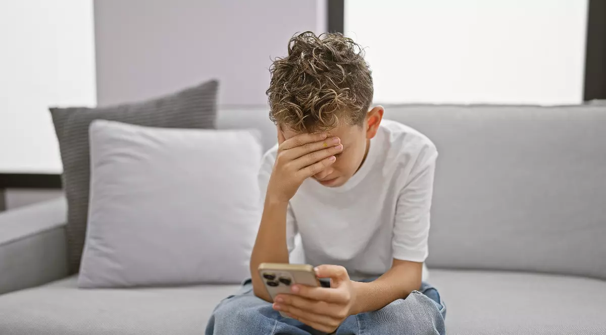 Imaginea prezintă un tânăr stând pe o canapea, ținând telefonul în mână și acoperindu-și fața cu cealaltă mână, semn de stres sau îngrijorare. Postura și expresia sugerează o stare de anxietate, posibil legată de conținutul pe care îl vede pe ecran.