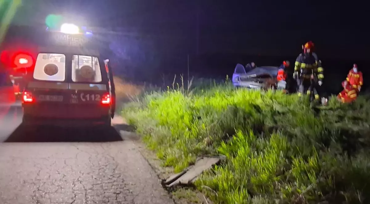 Imaginea surprinde intervenția echipajelor de urgență la locul unui accident rutier petrecut pe timp de noapte, pe marginea unui drum. În apropiere se observă o mașină avariată, iar pompierii și paramedicii acționează pentru gestionarea situației.