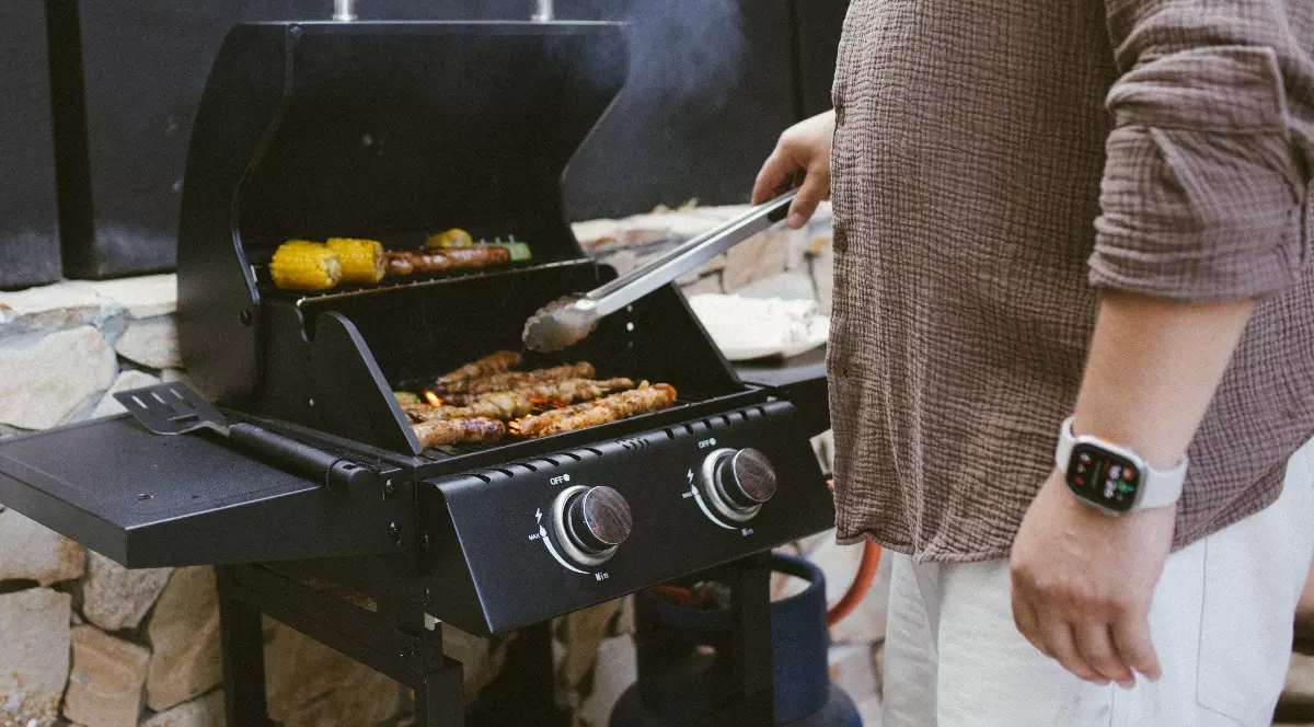 O persoană pregătește carne pe un grătar în aer liber, în timp ce fumul se ridică deasupra. Imaginea surprinde atmosfera relaxată a unei zile de primăvară, specifică ieșirilor de 1 Mai.